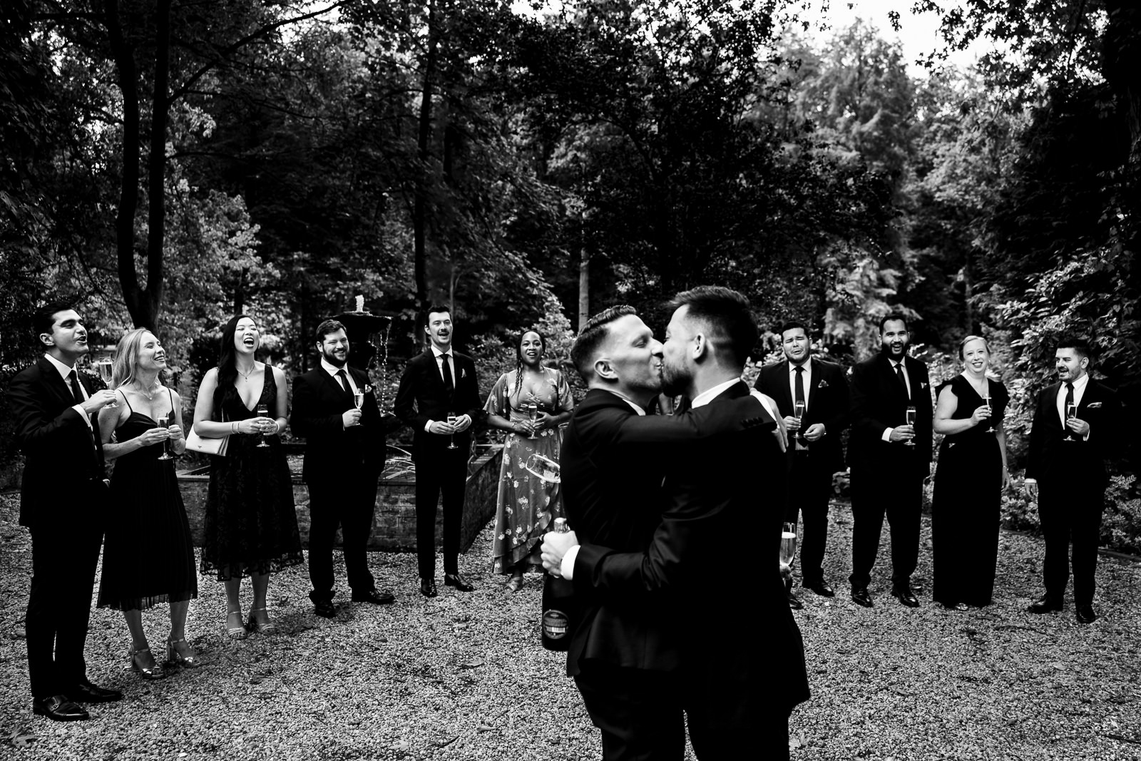 Two grooms kissing during same sex first look with bridal party watching